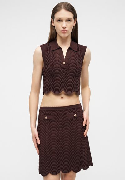 Femme portant un haut court sans manches en maille marron foncé et une jupe assortie jusqu'aux genoux avec des motifs ondulés et des boutons décoratifs.