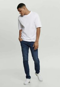 Λευκό βαμβακερό T-shirt με κοντά μανίκια, συνδυασμένο με μπλε τζιν παντελόνι και λευκά αθλητικά παπούτσια. Συνολικά, είναι ένα casual ντύσιμο με άνετη εφαρμογή.