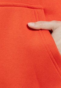 Tessuto arancione brillante con una superficie liscia e una cucitura della tasca ben visibile. Una mano è posizionata vicino all'apertura della tasca, evidenziando l'accessibilità.