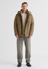 Braune isolierte Jacke mit gefütterter Kapuze, verschlossen mit Reißverschlüssen und Knöpfen, getragen über einem hellgrauen Langarmshirt und grauen Jeans.