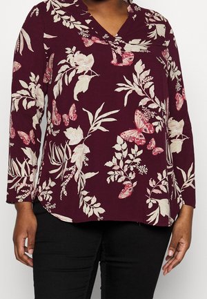 Blusa de manga larga de color púrpura oscuro con estampados blancos de flores y mariposas rosas, combinada con pantalones negros, mostrada desde el cuello hasta las caderas.