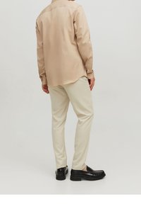 Beige overhemd met lange mouwen en een gladde textuur, gecombineerd met crème-kleurige broek en zwarte loafers. Het overhemd heeft een kraag en manchetten met knopen.