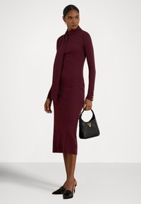 Μακρυμάνικο μπορντό φόρεμα με ψηλό γιακά και λεπτομέρεια με μαντήλι, συμπληρωμένο με μαύρη τσάντα με χρυσή λεπτομέρεια και μαύρα παπούτσια με τακούνι.