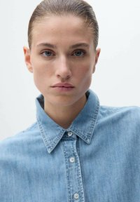 Helleblaue Jeanshemd mit spitzem Kragen, Knopfleiste vorne und einer weichen, glatten Textur. Die Details umfassen eine subtile Waschung und Nähte.