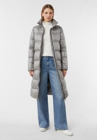 Lange, geïsoleerde pufferjas in lichtgrijs met een hoge kraag, ritssluiting en een gewatteerd ontwerp. Gecombineerd met wijde blauwe jeans en witte sneakers.