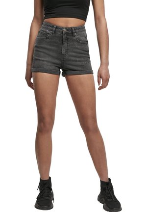 Nő fekete, rövidített tank toppal, magas derekú, sötétszürke farmer rövidnadrággal, felhajtott szegéllyel és fekete sportcipővel, fehér háttér előtt áll.