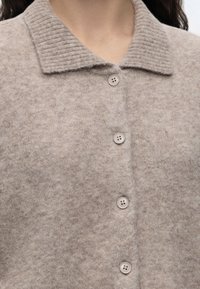 Gros plan d’un cardigan beige en laine à boutons avec un col côtelé porté par une personne aux cheveux foncés.