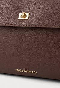 Smeđa kožna torba s teksturiranom površinom, zlatnim zatvaranjem i ugraviranim logotipom na dnu, koja ima elegantan pravokutni dizajn.