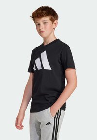 T-shirt de sport noir avec un grand logo blanc en diagonale sur le devant. Manches courtes, col rond, porté avec un pantalon de jogging gris à rayures.