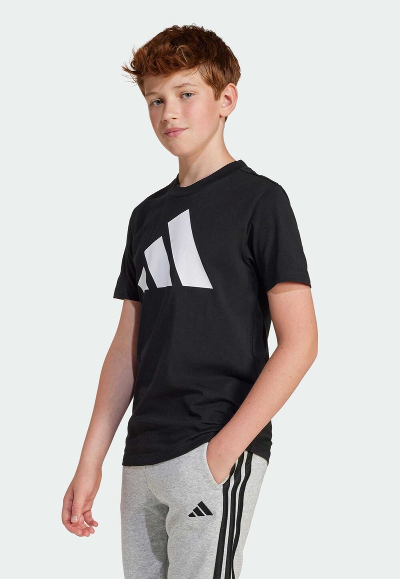 T-shirt de sport noir avec un grand logo blanc en diagonale sur le devant. Manches courtes, col rond, porté avec un pantalon de jogging gris à rayures.