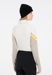 Langärmliges Sportshirt mit hohem Kragen, beigen Ärmeln, cremefarbenem Körper und gelben Chevron-Akzenten. Kombiniert mit schwarzen Sporthosen.