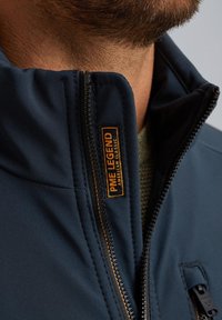 Dunkelblaues Zip-Up-Jackett aus strukturiertem Stoff, mit einem orangefarbenen Logo-Patch und einem hohen Kragen mit glattem Reißverschluss-Detail.
