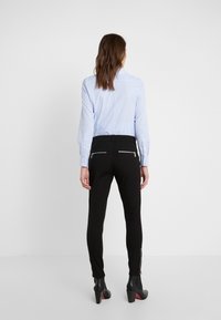 Pantalones negros ajustados con bolsillos con cremallera lateral, combinados con una camisa de botones de color azul claro, vistos desde la parte de atrás. Calzado negro de tacón.