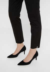 Svarta mocka högklackade skor med spetsiga tår, visade tillsammans med åtsittande svarta byxor med en sidoklyvning vid fållen.