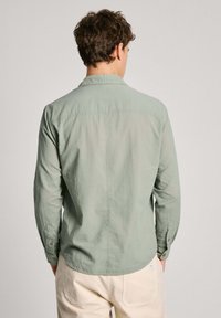 Camicia a maniche lunghe verde chiaro con una trama sottile, polsi con bottoni e orlo curvo, mostrata da dietro.