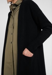 Svart stickad cardigan med långa ärmar, ribbade manschetter och sidofickor, kombinerad med en olivgrön skjorta med knappar under.