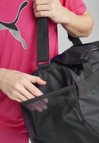 Black gym bag with a mesh pocket; top handles and textured fabric. A hand reaches into the bag, against a light background.