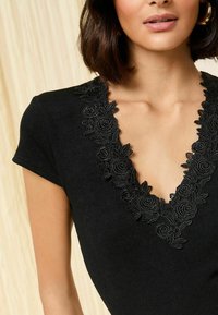 Top nero con scollatura a V decorato con intricati ricami floreali neri. Le maniche corte e la texture a costine arricchiscono il design.