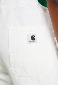 Des salopettes en coton blanc présentent une grande poche arrière avec une étiquette grise affichant un logo noir. Le tissu a une texture lisse.