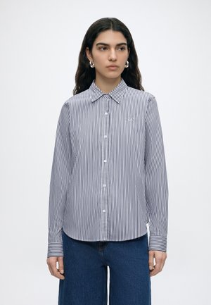 Frau mit langen dunklen Haaren, die silberne Creolen, ein weißes Hemd mit schwarzen vertikalen Streifen und Knöpfen sowie blaue Jeans trägt.