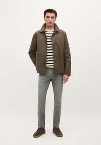 Olivgrüne Jacke mit Kragen, Knopfverschluss und großer Brusttasche, kombiniert mit einem gestreiften Pullover, hellen Jeans und braunen Schuhen.