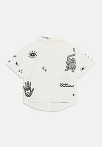 Kortermet skjorte med krage i lys kremfarge, som har svarte grafiske trykk av en tiger, bølger, hender og ulike symboler i hele skjorten.