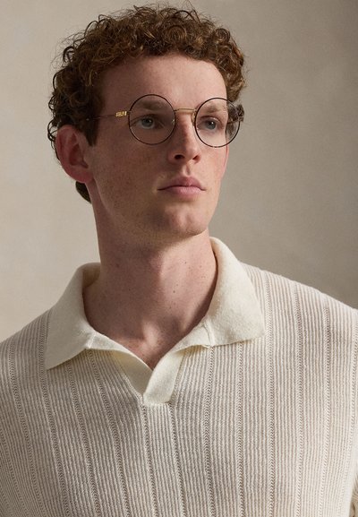 Νέος άνδρας με σγουρά κόκκινα μαλλιά και στρογγυλά γυαλιά, που φοράει πουλόβερ πόλο σε εκρού χρώμα με υφή πλέξης, κοιτάζοντας ελαφρώς προς το πλάι.
