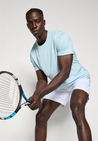 Light blue athletic t-shirt with short sleeves, paired with white shorts. The player holds a tennis racket, preparing to serve.
