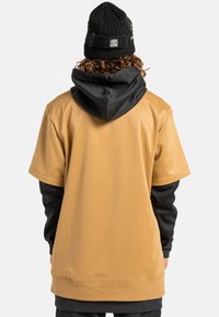 Beżowa bluzka z krótkim rękawem i czarnym kapturem, wykonana z gładkiego materiału. Posiada luźny krój oraz prążkowane akcenty na kapturze i rękawach.