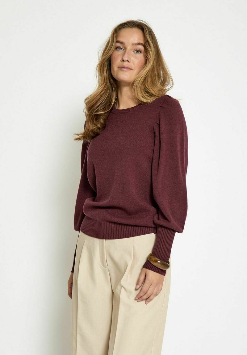 Burgundy knit sweater with ribbed cuffs and hem, slightly puffed sleeves, worn with light beige trousers and a chunky bracelet.