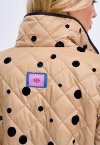 Béžová prošívaná bunda s velkými černými puntíky, vysokým límcem a fialovou vyšívanou nášivkou s okem a textem.