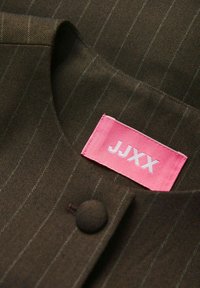 Brązowa tkanina w wzór rybiej ości z pionowymi paskami, z różową metką z białym tekstem "JJXX" i małym czarnym guzikiem.