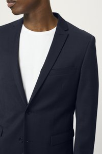 Námornícky blazer vyrobený z textúrovanej tkaniny, s klopou, dvoma gombíkmi a náprsníkom, položený na bielej košeli.
