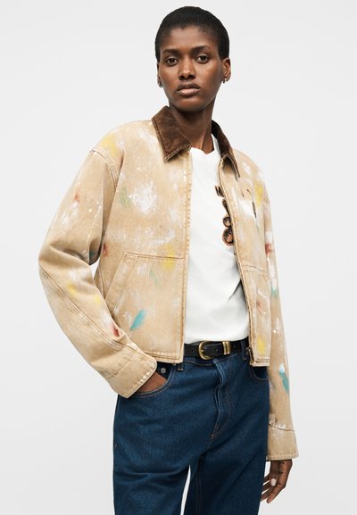 Γυναίκα που φοράει μια μπεζ μπουφάν με κηλίδες μπογιάς και καφέ γιακά, λευκό μπλουζάκι με σχέδιο, μπλε τζιν και μαύρη ζώνη, στεκόμενη με το χέρι στην τσέπη.