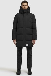 Cappotto imbacuccato nero con cappuccio, caratterizzato da una trama trapuntata, tasche con cerniera e un emblema sull'orlo. Indossato con jeans scuri e stivali.