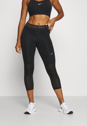 Vrouw staat in zwarte sportbeha en capri-leggings met Nike-logo's, witte sneakers, hand op de heup, tegen een effen witte achtergrond.