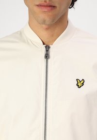 Lichtbeige bomberjack met een rits aan de voorkant, geribbelde kraag en een zwart geborduurd logo van een vogel op de linkerkant van de borst. Glad textuur.