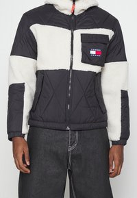 Padded jacket s černým exteriérem a bílými fleecovými akcenty. Obsahuje zipový uzávěr, kapuci a náprsní kapsu s detailem loga.