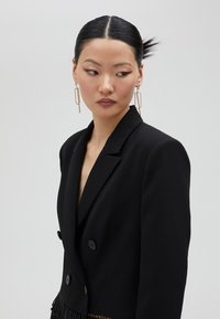 Svart dubbelknäppt kavaj med en texturerad frans vid fållen. Har två stora svarta knappar och en djup V-ringning.