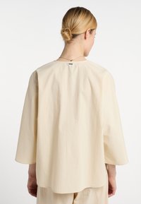 Beige, lockere Bluse mit Dreiviertelärmeln, rundem Ausschnitt und subtiler Textur. Enthält ein kleines Etikett auf der Rückseite.