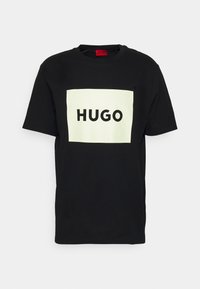 Černé tričko vyrobené z bavlny, se velkým bílým obdélníkovým logem s nápisem "HUGO" v tučných černých písmenkách na přední straně. Krátké rukávy.