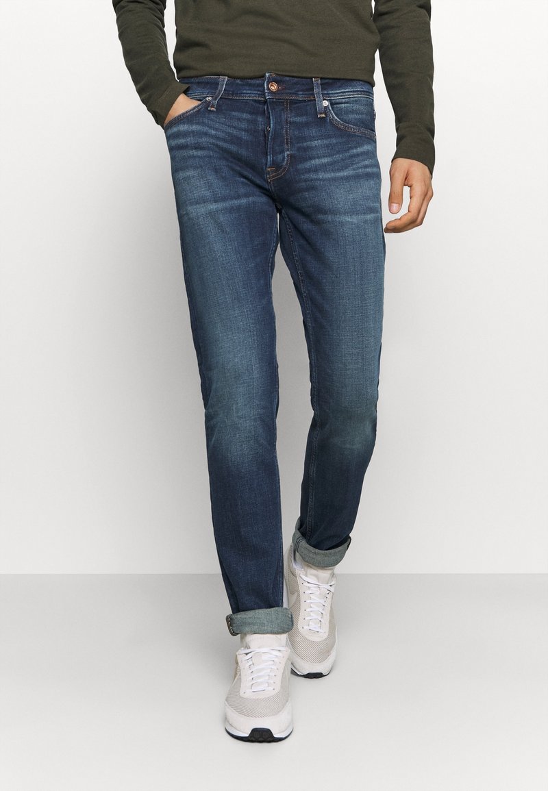 Mörkblå denimjeans med smal passform, med orange sömmar, främre fickor och uppvikta muddar. Kombinerade med ljusa sneakers.