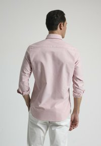 Camisa de manga larga rosa claro con puños doblados, yugo trasero y dobladillo curvado, combinada con pantalones blancos. Textura de tela suave.