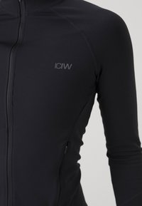 Melns sporta jaka ar rāvējslēdzēju, izgatavota no elastīga auduma. Tai ir priekšējais rāvējslēdzējs un neliels logo kreisajā krūtīs, ar pieguļošām garām piedurknēm.