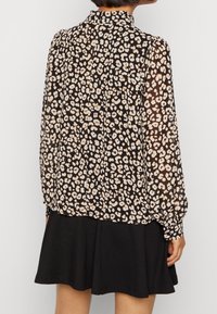 Durchsichtige schwarze Bluse mit beigen Leopardenflecken, hohem Kragen, plissierten Ärmeln und knopfartigen Manschetten, kombiniert mit einem schwarzen, ausgestellten Rock.