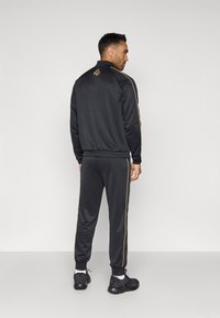 Čierny teplákový súprava so zlatými bočnými pruhmi, vyrobená z hladkého materiálu. Obsahuje malý vyšívaný detail na hornej časti chrbta. Zodpovedajúce nohavice a topánky.