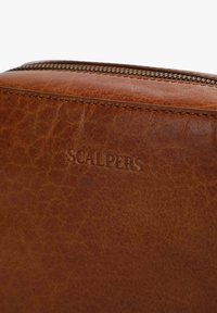 Brown leather bag corner with embossed "SCALPERS" logo, featuring a zipper detail and visible textured grain in the material.
