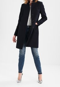 Granatowy długi płaszcz o prostym kroju, z kołnierzem i trzema guzikami z przodu. W zestawieniu z jasnoniebieskimi dżinsami i srebrnymi szpilkami.