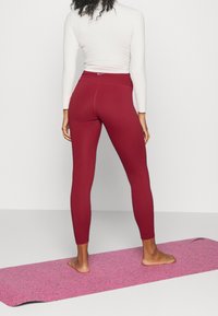 Kvinde stående barfodet på en pink træningsmåtte, iført røde leggings og en hvid langærmet top, vendt væk fra kameraet.