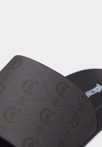 Μαύρο σανδάλι με φαρδιά υφή λουρί διακοσμημένο με διακριτικό επαναλαμβανόμενο λογότυπο "D" και αναπαυτικό πέλμα ορατό στο φόντο.